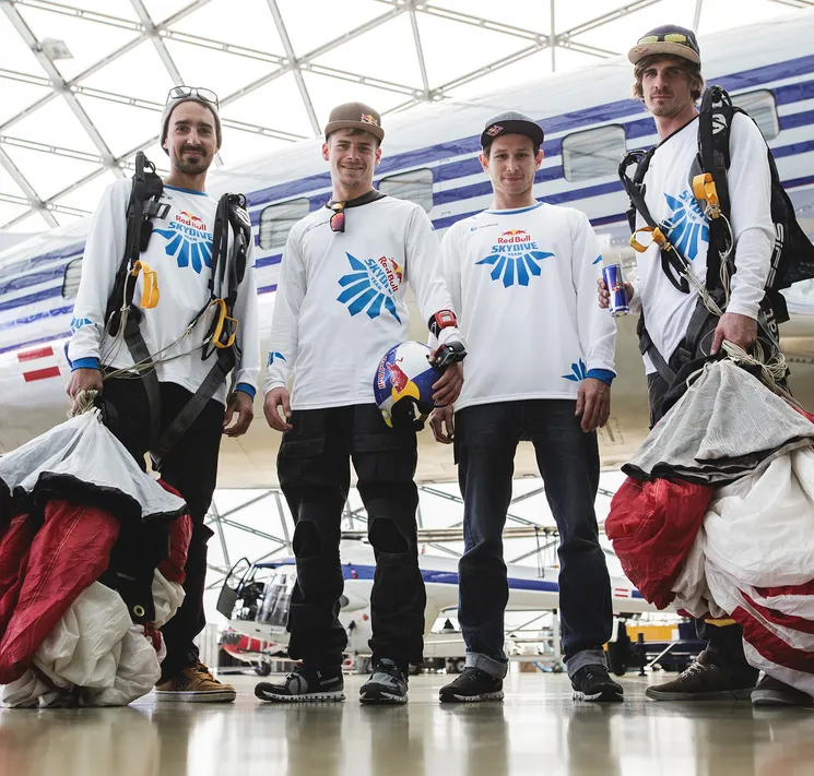 Vier Männer in weißen, langärmeligen Sportshirts mit dem Logo der Red Bull Skydive Team stehen in einer Halle vor einem Flugzeug.