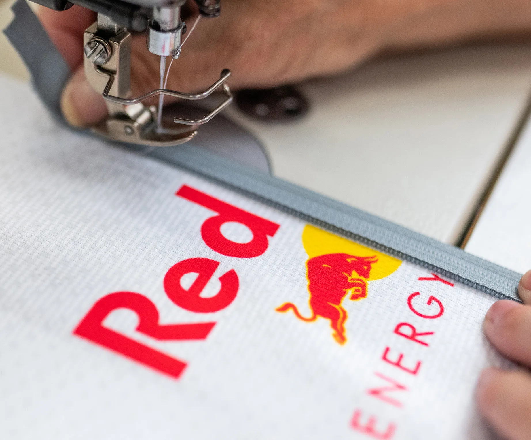 Eine Nähmaschine näht einen grauen Reißverschluss an ein weißes Sportbekleidungsstück mit dem Red Bull-Logo.
