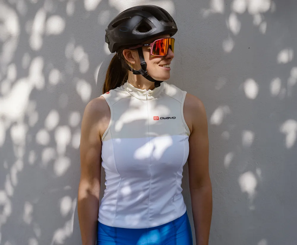 Eine Frau in einem weißen, ärmellosen Radtrikot mit dem Logo von owayo, einem Fahrradhelm und Sonnenbrille, lehnt an einer Wand mit Blattenschatten.