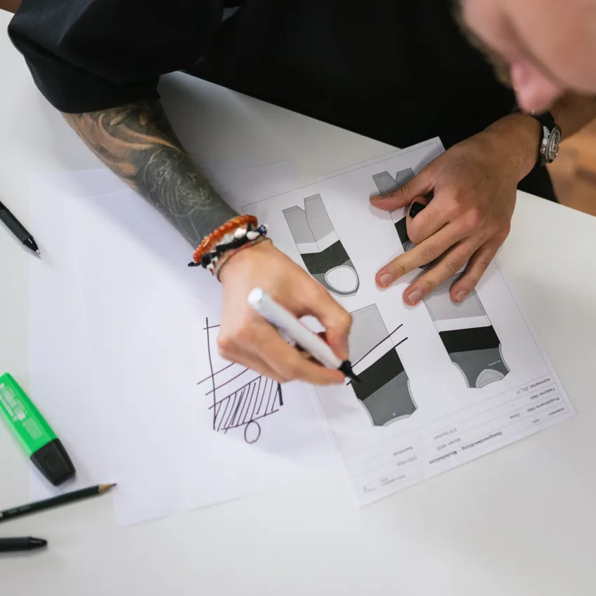 Eine Person mit einem tätowierten Arm skizziert Entwürfe für Sportbekleidung auf Papier, umgeben von Schreibutensilien.