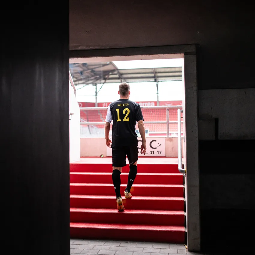 Ein Fußballspieler in einem schwarzen Trikot mit der Nummer 12 geht die roten Stufen eines Stadions hinauf.