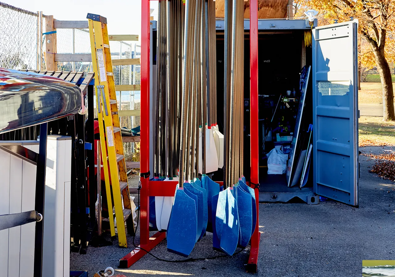 Ein Bild von einem Bootslager mit Ruderblättern in verschiedenen Farben, einem gelben Leiter und einem offenen Container im Hintergrund.