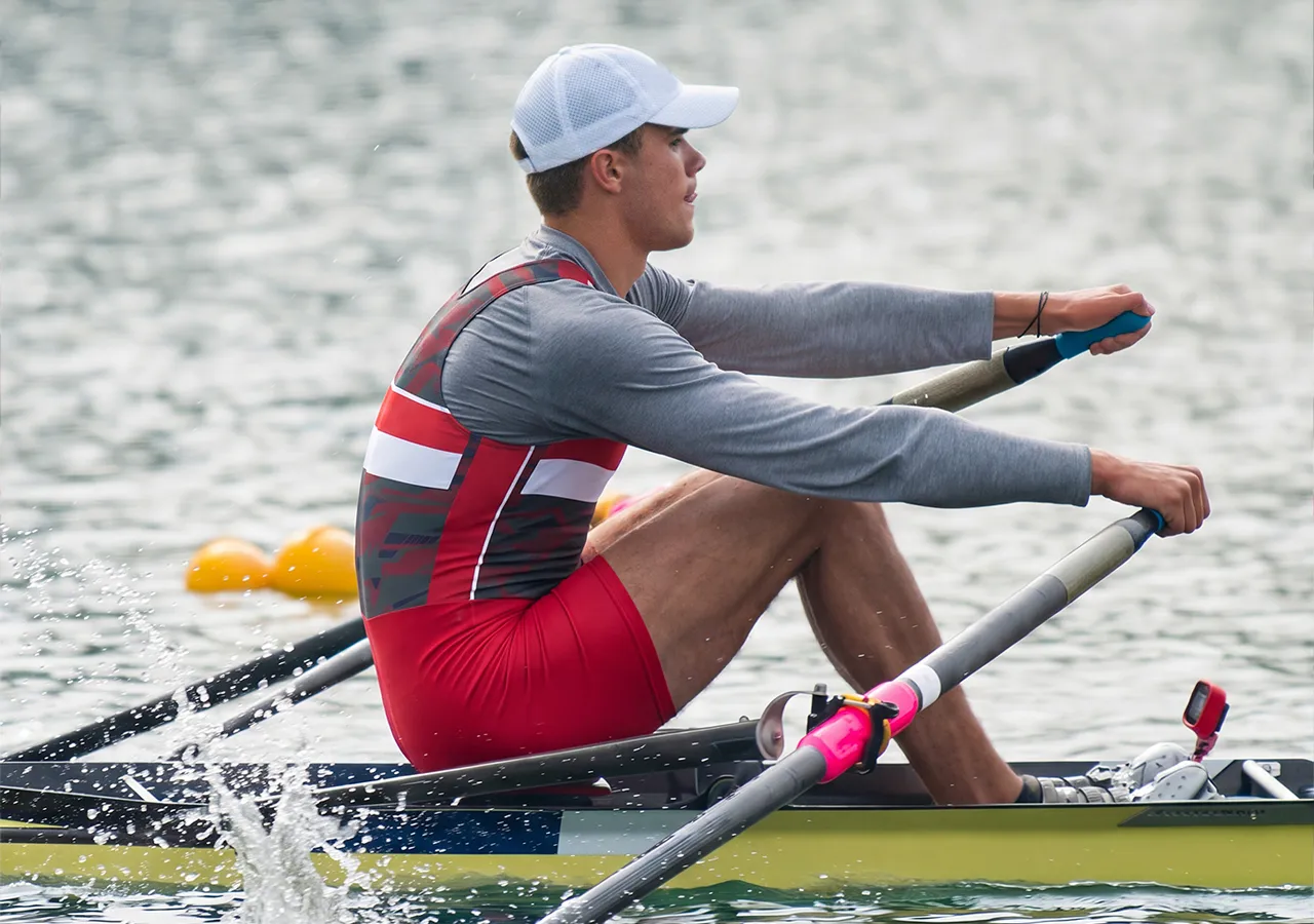 Ein Ruderer in einem rot-grauen Sportoutfit rudert auf einem Gewässer.