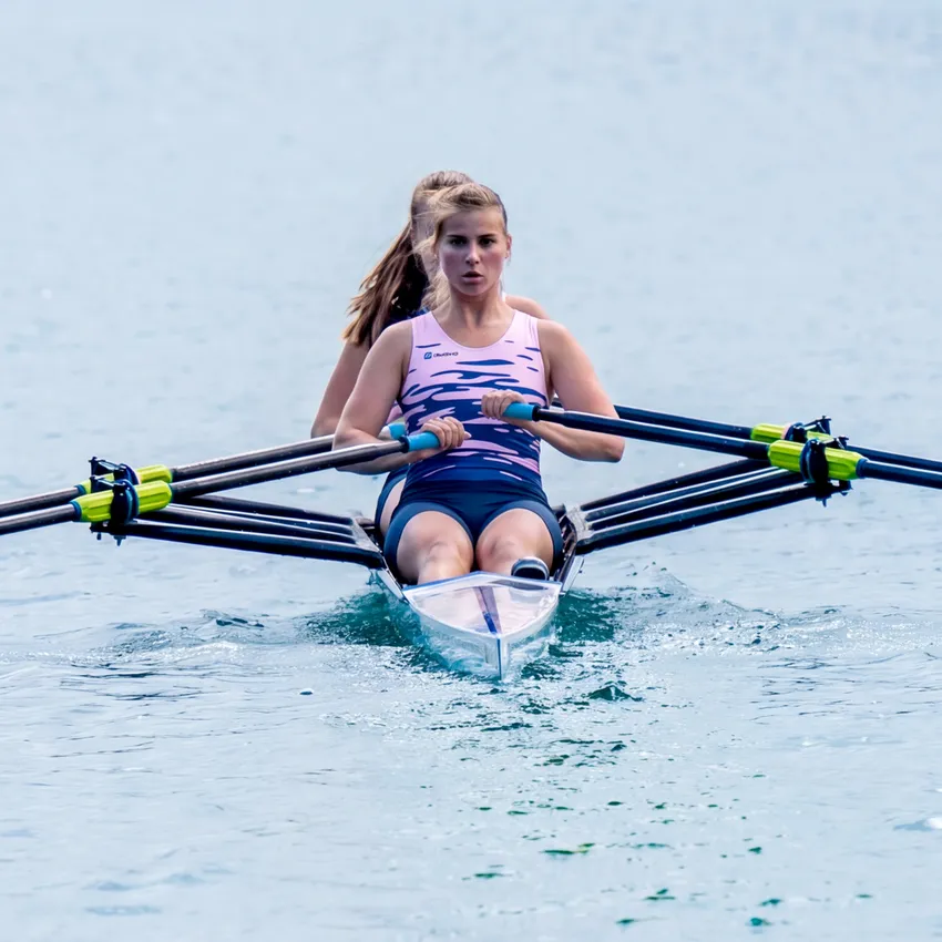 Zwei Ruderinnen in einem Boot, die auf ruhigem Wasser rudern, wobei die vordere Rudererin ein pinkfarbenes Sportoutfit trägt.