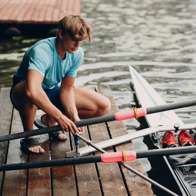 Ein junger Mann in einem blauen Sportshirt sitzt auf einem Steg und montiert Ruder für ein Boot, während im Hintergrund Wasser und ein Boot zu sehen sind.