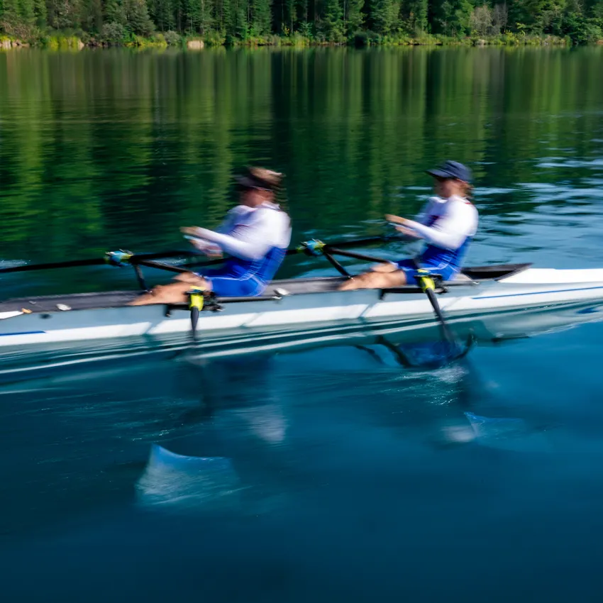 Zwei Ruderer in blauer Sportbekleidung, die mit kraftvollen Bewegungen über einen ruhigen See rudern.