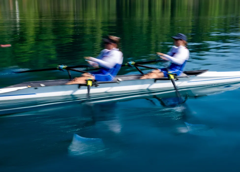 Zwei Ruderer in blauer Sportkleidung rudern synchron auf einem ruhigen Gewässer.