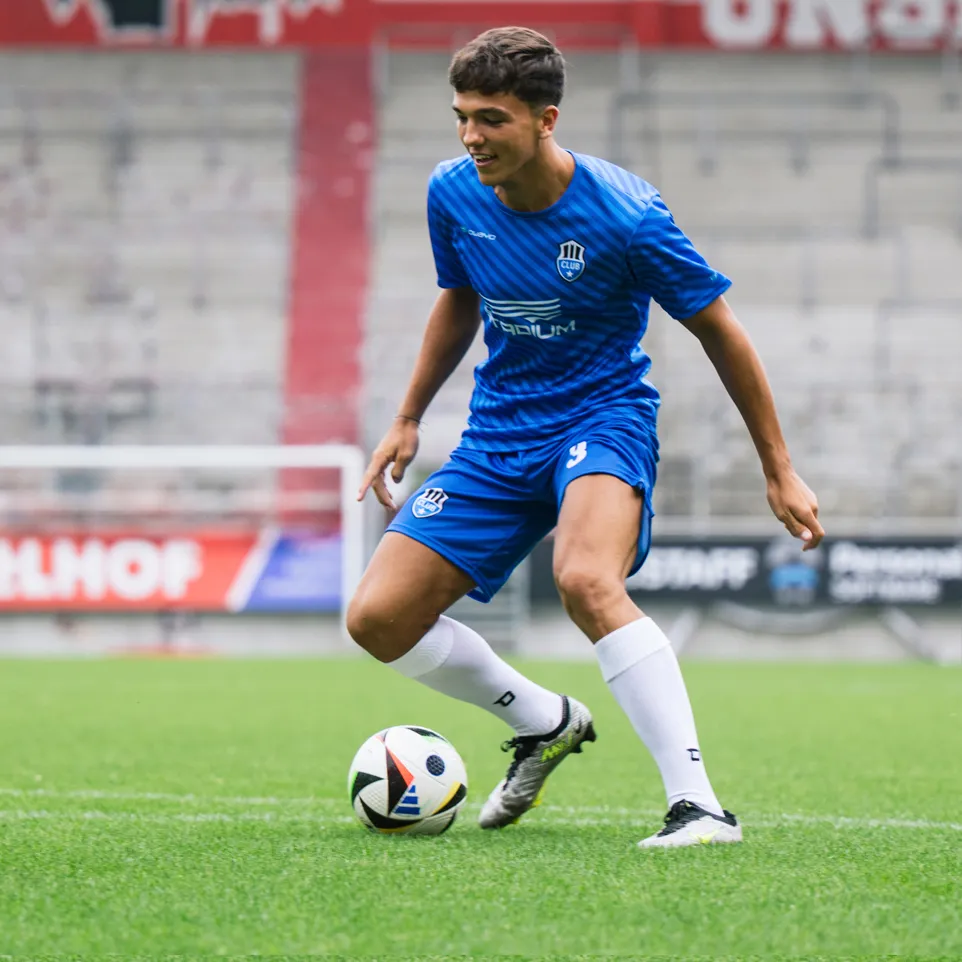 Ein junger Fußballspieler in einem blauen Trikot und blauen Shorts mit einem Logo auf der Brust und der Nummer 3 auf der Hose dribbelt einen Fußball auf einem grünen Rasenplatz.