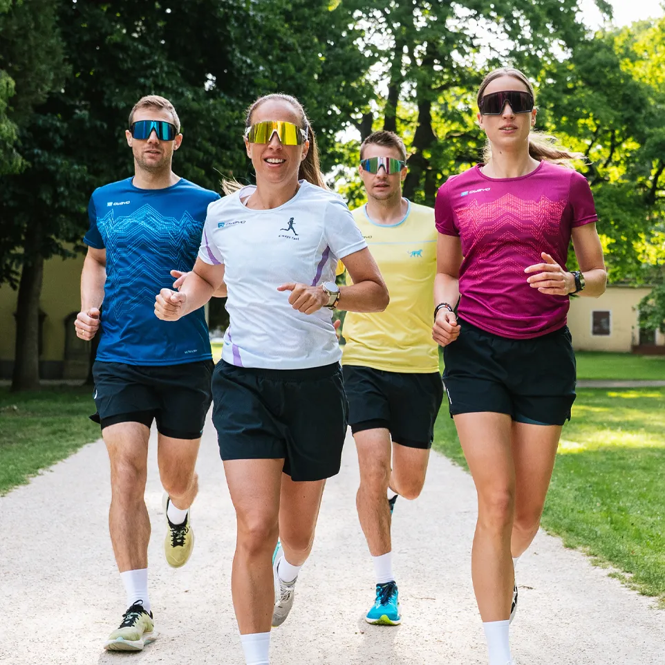 Vier Läufer in bunten Sportshirts und Shorts joggen auf einem Weg in einem Park.