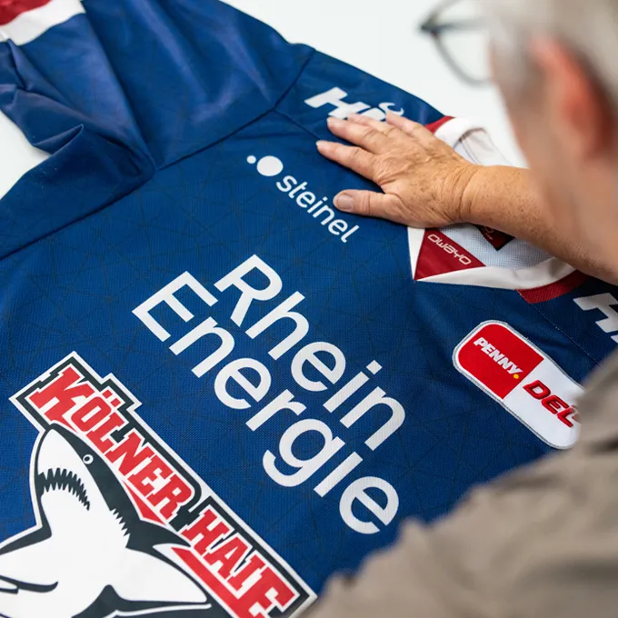Eine Person prüft ein blaues, maßgeschneidertes Sporttrikot von owayo mit verschiedenen Sponsorenlogos, darunter 'Rhein Energie' und 'Kölner Haie'.
