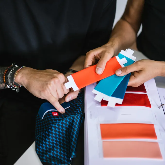 Zwei Personen wählen Farbproben für maßgeschneiderte Sportbekleidung aus, wobei ein blauer Stoff mit Muster und verschiedene Farbvarianten zu sehen sind.